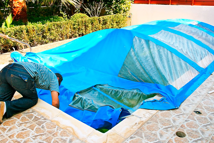 A man covers a swimming pool with a blue tarp, preparing for maintenance in an Austin backyard.